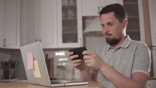 Adulto bonito homem está jogando em um smartphone em casa, usando fones de ouvido — Vídeo de Stock