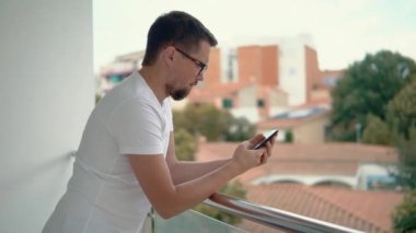 Yetişkin sakallı adam balkon korkulukları üzerinde eğilerek telefonla mesaj okuyor