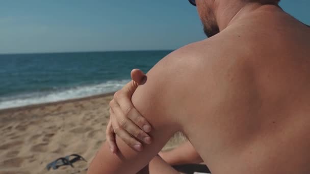 Man is applying sunscreen cream in his skin, sitting on a beach in summer day — Stock Video
