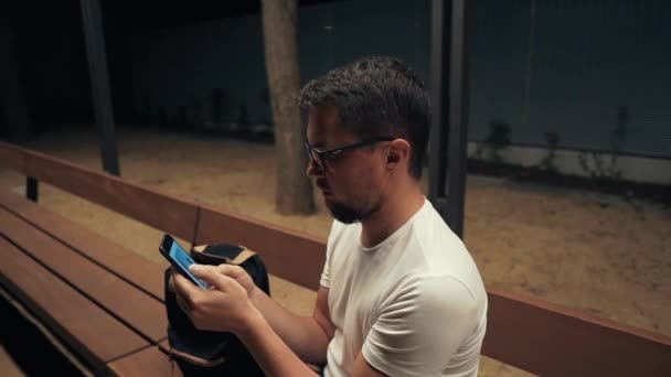 Adult man is watching pictures in mobile phone, sits alone in evening outdoors — Stock Video