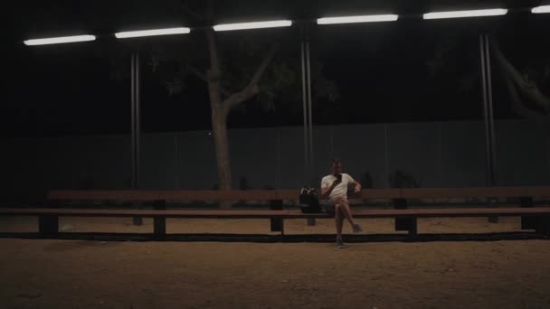 Горожанин сидит один в ночное время в парке под уличными фонарями — стоковое видео