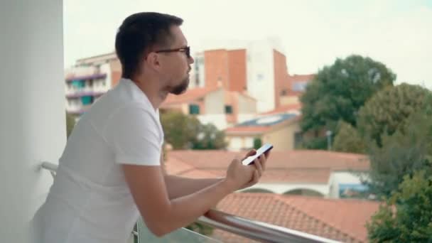 Młody człowiek jest przesuwając palcem na ekranie dotykowym telefonu na balkonie mieszkania w małym mieście — Wideo stockowe