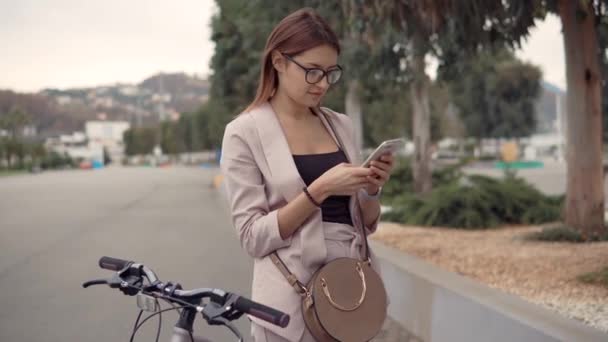 Mujer morena joven está escribiendo sms en el teléfono móvil, de pie cerca de una bicicleta — Vídeos de Stock