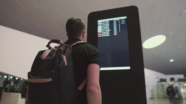 Νέος άντρας με σακίδιο παρακολουθεί ηλεκτρονικά δρομολόγια στην αίθουσα αεροδρομίου — Αρχείο Βίντεο