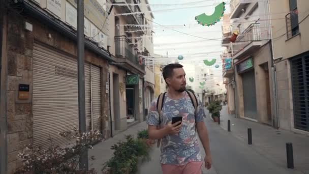 Ο νεαρός άνθρωπος περπατά στο δρόμο της πόλης, κρατώντας το τηλέφωνο στο χέρι, κοιτάζοντας στην οθόνη — Αρχείο Βίντεο