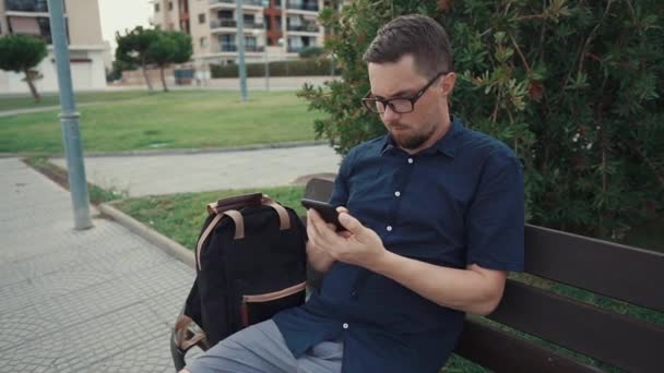 Hombre serio está sentado en un banco en el callejón en el parque de la ciudad, escribiendo en el teléfono móvil — Vídeos de Stock