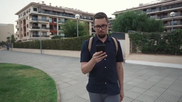 Vrolijke man loopt in vierkant in de zomer dag, op zoek op het scherm van zijn mobiele — Stockvideo