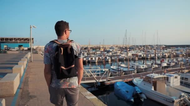 Мужской турист наслаждается прогулкой по морскому пирсу с яхтами в солнечное утро — стоковое видео