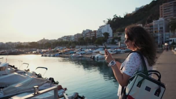 Townswoman is standing alone on sea port in town, photographing by smartphone — Stock Video