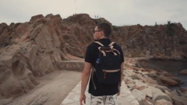 Erkek gezgin gündüz deniz kıyısında taş yolu boyunca gidiyor, arka görünüm