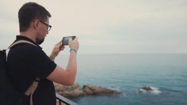 Erkek turist akıllı telefon kamera kullanarak, alacakaranlık içinde deniz manzarası fotoğraflama