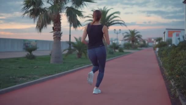 Mujer delgada está trotando en el terraplén de la ciudad a lo largo de pequeñas casas, mantenerse en forma — Vídeos de Stock