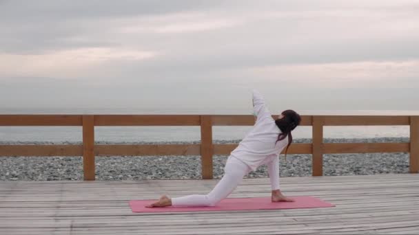 Женщина меняет асаны йоги, сидя на полу, на фоне моря — стоковое видео