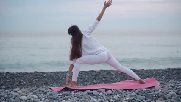 Woman is performing yoga asanas on pebble beach of sea in cloudy day, back view — Stock Video