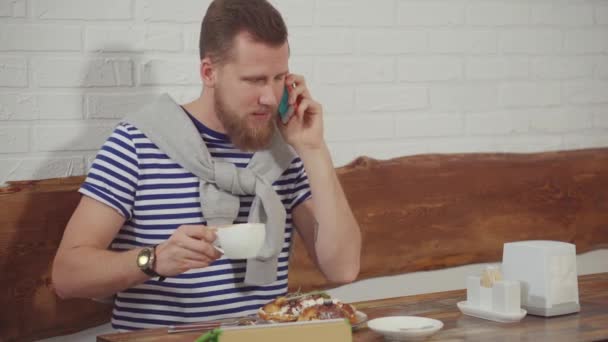 Homem está conversando por telefone sentado no café e beber café, sorrindo alegremente — Vídeo de Stock