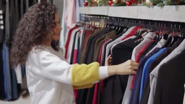 Cliente femenino está de compras en la tienda de ropa, sonriendo y mirando en los bastidores — Vídeos de Stock