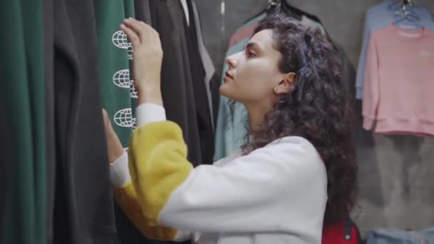 Adolescente chica está viendo de moda sudaderas en la tienda con ropa juvenil — Vídeos de Stock