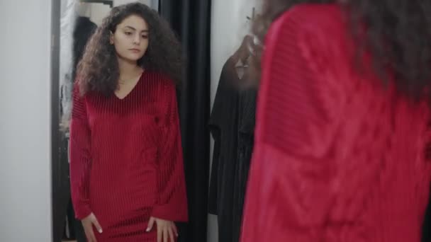 Μελαχρινή γυναίκα κοιτάζει στον καθρέφτη στην αλλαγή δωματίου στο κατάστημα, προσπαθώντας νέο φόρεμα — Αρχείο Βίντεο