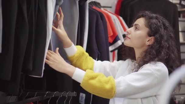 Женщина-клиент изучает ценники на спортивные футболки в магазине одежды — стоковое видео