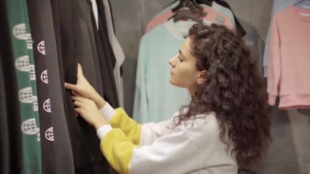 Женщина-клиент просматривает толстовки с узорами в магазине одежды — стоковое видео