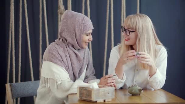 Muslim woman is talking cheerfully with her blonde friend in cafe, drinking tea — Stock Video
