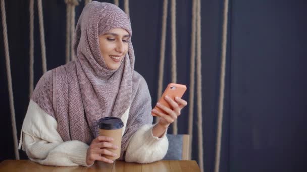 Volwassen vrolijke moslimvrouw is het lezen van SMS in mobiele telefoon en glimlachend in café — Stockvideo