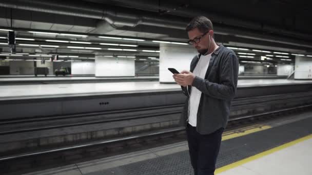 Чоловік стоїть біля підземного поїзда і чекає, використовуючи навушники — стокове відео