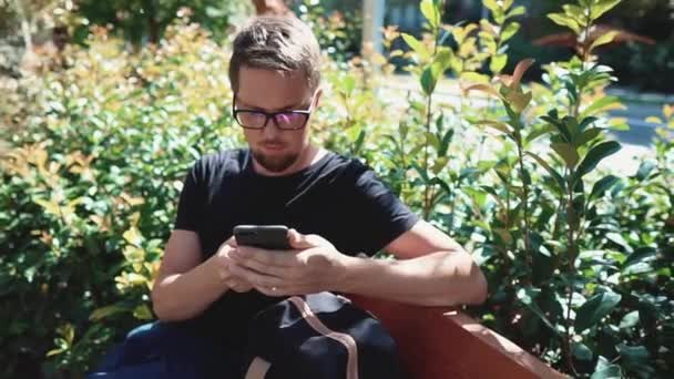 Homem está navegando redes sociais por seu smartphone ao ar livre no parque, sente-se no banco — Vídeo de Stock