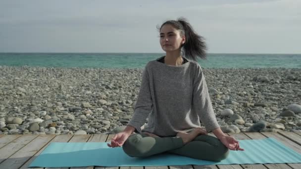 Женщина спокойно отдыхает на берегу моря, делая йогу асана, сидя на полу — стоковое видео