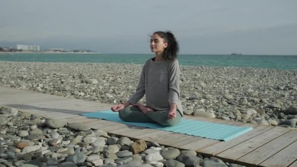 Relajada chica deportiva está sentado en el camino de madera cerca del mar y meditando — Vídeos de Stock
