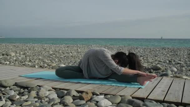 Спортсменка растягивает свое тело, сидя на деревянной дорожке на берегу моря. — стоковое видео