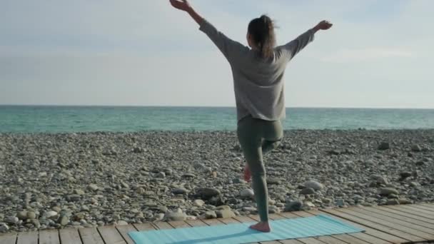 Μελαχρινή γυναίκα τεντώνει τα χέρια πάνω, στέκεται σε ένα πόδι στη θάλασσα ακτή, πίσω όψη — Αρχείο Βίντεο