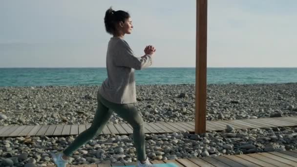 Young sporty woman is training outdoors near empty sea coast, squatting — 비디오