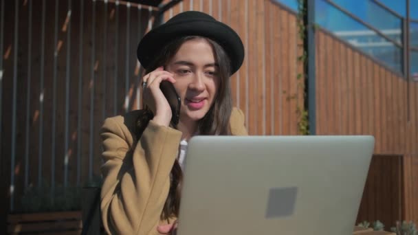 Mujer está llamando por teléfono y mirando en la pantalla de la computadora portátil pensativamente al aire libre — Vídeos de Stock
