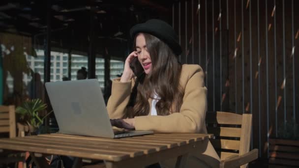 Trabajadora freelancer joven trabaja con laptop y habla por teléfono en la cafetería — Vídeo de stock