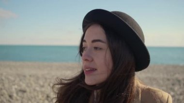 Kadın etrafına bakıyor güneşli sahilde oturuyor, saçlarına dokunuyor, yakın plan portre yapıyor.