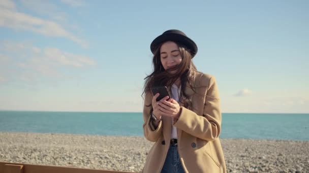 Молодая стильная девушка чатится в социальных сетях в смартфоне на берегу океана — стоковое видео