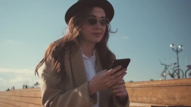 Brunett tjej använder smartphone, sitter utomhus i soliga dagar, människor bakom — Stockvideo