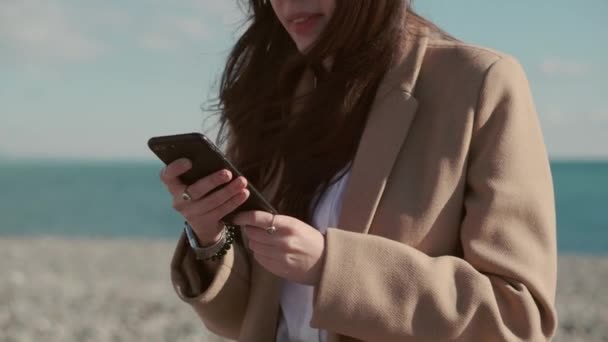 Joyful brunette woman is tapping on display of smartphone on beach, close-up — Stock Video