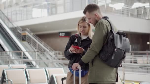 Молода пара перевіряє інформацію в смартфоні, що стоїть в залі аеропорту — стокове відео