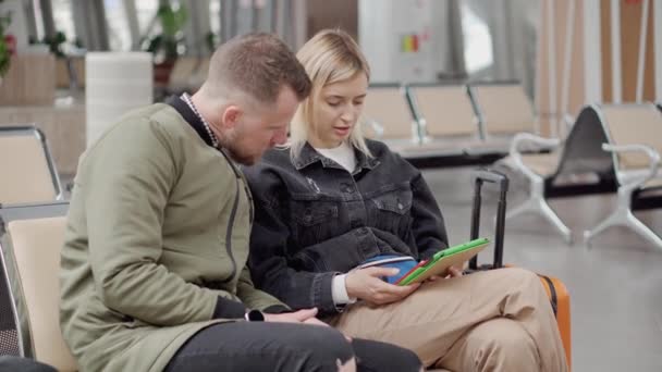 Jonge echtgenoten zitten samen in de vertrekhal en kijken op tablet — Stockvideo