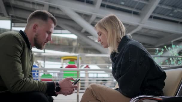 Mann und Frau sitzen in der Abflughalle und schauen auf den Bildschirm ihres Smartphones — Stockvideo