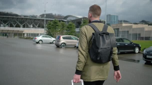 Мужчина путешественник ходит с чемоданом над парковкой станции, вид сзади — стоковое видео