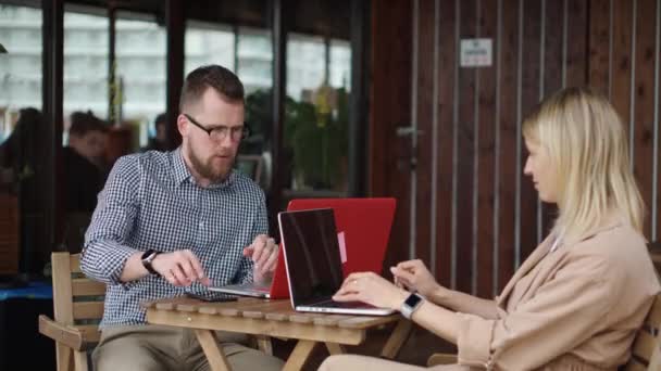 Молодая леди и красивый парень сидят за столом в открытом кафе, работают с ноутбуками — стоковое видео