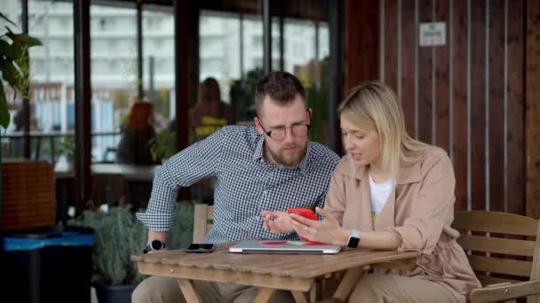 Dos amigos hombres y mujeres están sentados en la mesa en la cafetería, utilizando teléfonos inteligentes — Vídeos de Stock