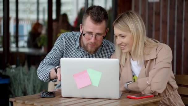 Les jeunes conjoints regardent l'écran d'ordinateur portable dans le café en terrasse ouverte, discuter — Video