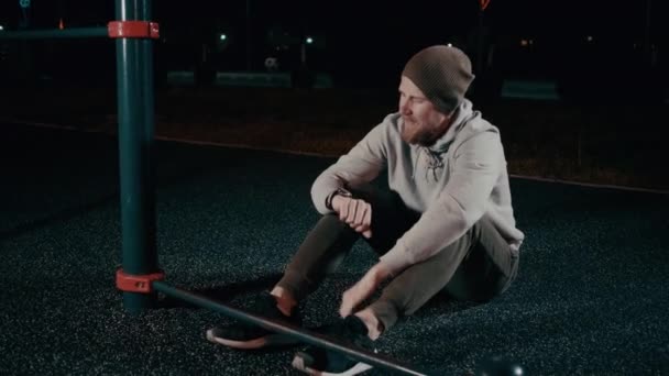 Втомлений спортсмен відпочиває після тренування в парку вночі, сидячи на землі — стокове відео