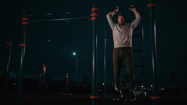Sportivní muž zvedá nohy zavěšené na vodorovném baru venku v noci. — Stock video