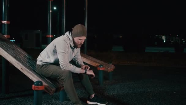 El hombre está sentado en el campo de deportes en el parque por la noche y mirando en smartwatch — Vídeos de Stock