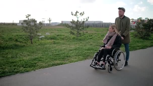 Man is rolling wheelchair with young blonde woman, walking together in park — Stock Video